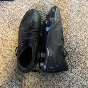 Svarta Adidas Predator fotbollsskor - Säljer ett par helsvarta Adidas Predator fotbollsskor med snörning och mönstrad ovandel. Skorna har en modern design med greppig sula för bästa fäste på planen. Perfekta för dig som vill sticka ut med stilren look på matchen. Använd 1 säsong, hade använt en till om det inte var för att jag fick för stora fötter. Storlek 38.