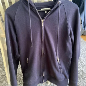 Suitsupply - Mörkblå ziphoodie från suitsupply. Storlek XS men sitter större (~S). I mycket gott skick förutom att pluppen på ena huvspännet har lossnat (se bild). Det är dock inget man tänker på överhuvudtaget. Nypris skulle jag tippa på ligger runt ~2000kr.