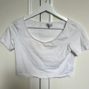Vit croppad topp från H&M Divided - Basic vit croppad topp från H&M Divided. Modellen har korta ärmar och en rundad, lite djupare halsringning. Tillverkad i mjuk bomull som känns skön mot huden. Perfekt att matcha med jeans eller kjol för en clean look.