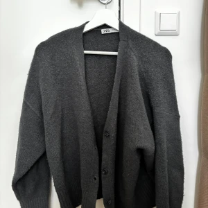 Mörkgrå kofta från Zara - Mysig mörkgrå kofta från Zara i storlek S. Koftan har en loose passform, djup v-ringning och knäppning framtill med tre knappar. Perfekt för lager-på-lager och chill dagar. Materialet känns mjukt och lite fluffigt, vilket gör den extra skön att bära. Är lite nopprig, köpte den på Vinted men har ej använt då den är för stor