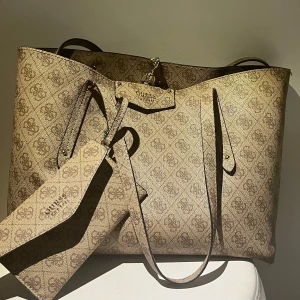 Beige shoppingväska från Guess - Snygg shoppingväska från Guess i beige med logomönster över hela väskan. Väskan har långa axelband, guldfärgade metalldetaljer och medföljande matchande kuvertväska. Tillverkad i syntetmaterial med Guess-logga framtill.