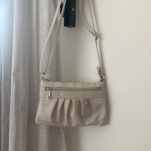 Beige axelväska med dragkedja - Snygg beige axelväska i mjukt material med justerbar rem. Väskan har ett veckat parti framtill och en praktisk dragkedja på framsidan och baksida för småsaker. Har även flera små fickor inuti. Perfekt storlek för det viktigaste och enkel att matcha till olika outfits.