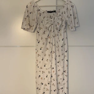 Vit blommig långklänning med snörning - Superfin långklänning från Lager 157 i vit med små blommor i beige och mörkblå. Klänningen har korta puffärmar, snörning framtill och en söt v-ringning. Materialet är mjukt och känns som bomull, perfekt för sommarens alla dagar.