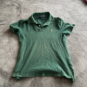 Grön pikétröja från Ralph Lauren - Klassisk grön pikétröja från Polo Ralph Lauren med korta ärmar och orange broderad logga på bröstet. Priset är diskuterbart, om du ha enögts frågor skriv