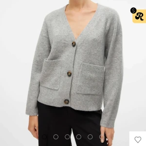 Grå kofta med stora knappar från Vero Moda - Mysig grå kofta med v-ringning och tre stora, mönstrade knappar framtill. Koftan har två rejäla fickor och ribbade detaljer vid ärmslut och nederkant. Perfekt för lager-på-lager och chill dagar. Helt ny aldrig använd!