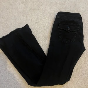 Svarta Joey Low Rise Flare jeans - Snygga svarta jeans från True Religion, modell Joey Low Rise Flare. De har låg midja och utsvängda ben. Fint skick inga lösa sömmar mm. Storlek 27