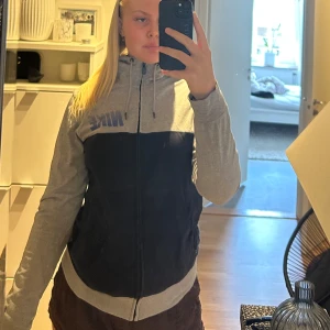 Nike kofta  - Jag säljer en jättefin kofta från Nike som är grå upptill samt på baksidan och ärmarna, svart nedtill och en blå Nike logga på bröstet. Koftan har även en dragkedja hela vägen och snörning i huvan. Priset kan även diskuteras! Denna kofta fungerar utmärkt för en promenad, träning eller bara till en vanlig outfit🥰