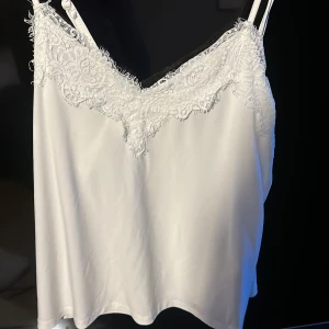 Vit spetslinnetopp från new yorkerXS - Supersöt vit linnetopp från new yorker i storlek XS. Toppen har tunna axelband och v-ringning med  spetsdetalj framtill och baktill. Materialet är glansigt och perfekt för att lyfta vilken outfit som helst.
