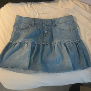 Blå jeanskjol med volang från edc - Supersöt ljusblå jeanskjol från edc med volang nertill och tre knappar framtill. Kjolen har klassiska jeansfickor och bälteshällor. Perfekt för dig som gillar denim och vill ha en chill och trendig look.