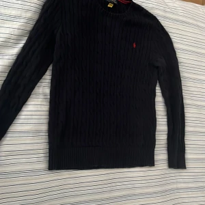 Svart kabelstickad tröja Polo Ralph Lauren - Svart kabelstickad tröja från Polo Ralph Lauren i storlek XL. Tröjan har ett klassiskt rött Polo-broderi på bröstet, rund hals och långa ärmar. Ribbad mudd vid ärmslut och nederkant. Perfekt för dig som gillar stilren och tidlös design. Skicka pm om ni har frågor köpt på NK om man undrar 