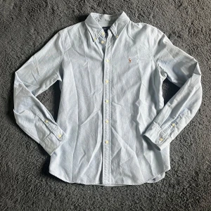 Ljusblå skjorta från Ralph Lauren - Snygg ljusblå skjorta från Ralph Lauren i storlek L, custom fit. Klassisk button-down krage, långärmad och med vita knappar framtill. Broderad logga på bröstet. Skjortan är gjord i mjuk bomull och har en stilren, normal passform. STORLEKEN på etiketten är L men skjortan motsvarar mer M heller S så kolla måtten noggrant ! (Jag skickar ej bild när jag bär plaggen )