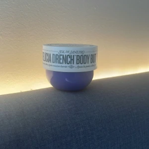 Sol de Janeiro Delicia Drench Body Butter - En burk Delicia Drench Body Butter från Sol de Janeiro i en snygg lila plastförpackning med vitt lock. Krämen är vit och har en rik, återfuktande formula som passar perfekt för torr hud. Doften är fräsch och burken är rund och kompakt. 