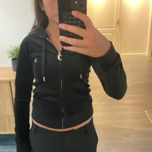 Svart Juicy Couture hoodie i sammet - Svart hoodie från Juicy Couture i mjuk sammet med glittrande detaljer. Dragkedja framtill med J-berlock, huva och snörning. På ryggen står det 'Juicy' i strass och framtill finns en liten JC logga