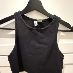 Svart croppad linnetopp H&M Divided - En svart croppad linnetopp från H&M Divided. Toppen har en enkel och stilren design med breda axelband och rund halsringning. Perfekt att matcha med höga jeans eller kjol för en chill look.