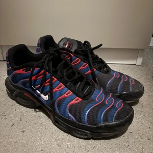 Nike Air Max Plus Tn blå/röd sneakers - Nike Air Max Plus Tn sneakers med svart mesh-överdel, blå och röda vågiga detaljer och klassisk Tn-logga på hälen. Snygg design med svarta snören och robust svart sula. Perfekt för dig som gillar streetstyle och vill sticka ut med färg och mönster.