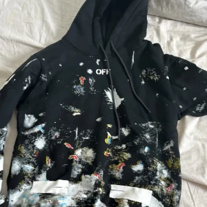 Svart hoodie från Off-White med färgstänk - Säljer en svart hoodie från Off-White med coola färgstänk i vitt, gult, blått och rött. Tröjan har huva med snörning och Off-White-logga på bröstet. Materialet är mjuk bomull och passformen är normal. Perfekt för dig som gillar streetwear och unik design.
