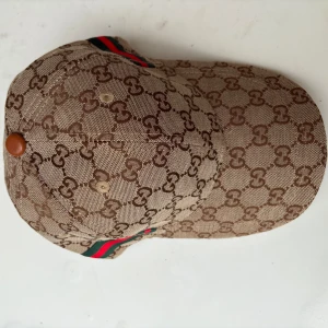 Gucci keps med GG-mönster och läderdetalj - Exklusiv keps från Gucci i beige och brunt med klassiskt GG-monogram över hela kepsen. Justerbar rem i brunt läder bak och ikoniska gröna och röda ränder på sidan. Tillverkad i merinoull och kashmirblandning, insida med Gucci-logga.