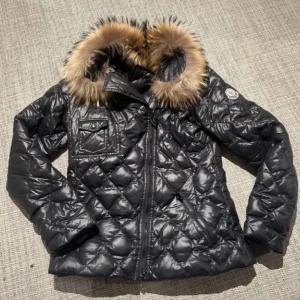 Svart Moncler dunjacka med päls (den är sönder och funkar inte att zippa” - Snygg svart dunjacka från Moncler med quiltad design och fluffig päls runt huvan. Jackan har dragkedja framtill, en bröstficka med knapp och Moncler-logga på ärmen. Perfekt för kalla dagar och har en cool, lyxig vibe.