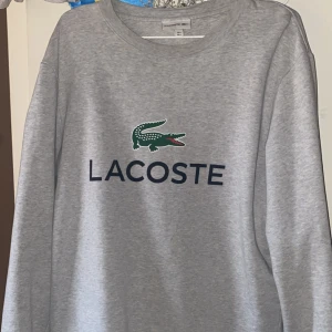 Grå sweatshirt från Lacoste - Snygg grå sweatshirt från Lacoste med klassisk krokodil-logga och stor text på bröstet. Tröjan har rund halsringning och långa ärmar, perfekt för en chill och stilren look. Tillverkad i mjuk bomullsmix som är skön att ha på sig hela dagen. Köpt för 1,500