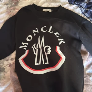 Svart sweatshirt från Moncler - Svart sweatshirt från Moncler med stort vitt och rött logotryck på bröstet och klassisk rund hals. Tröjan har långa ärmar och en broderad Moncler-patch på ena ärmen. I stl s/xs