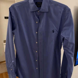 Blå skjorta från Polo Ralph Lauren - Snygg blå skjorta från Polo Ralph Lauren i slim fit-modell. Skjortan har klassisk krage, långa ärmar och knappar framtill. Broderad logga på bröstet ger en clean och stilren look. Perfekt för dig som gillar enkel men exklusiv stil.