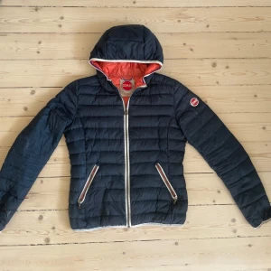 Mörkblå Colmar pufferjacka med huva - Snygg mörkblå pufferjacka från Colmar med orangefärgat foder och huva. Jackan har två dragkedjeförsedda fickor och tydliga logodetaljer på bröstet och ärmen. Perfekt för kyliga dagar och har en sportig vibe.