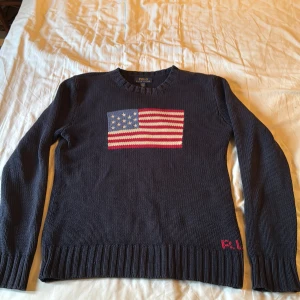 Mörkblå stickad tröja Polo Ralph Lauren, passar perfekt nu inför hösten - Säljer en mörkblå stickad tröja från Polo Ralph Lauren i storlek 14/16 vilket motsvarar xs. Tröjan har ett stort amerikanskt flaggmotiv på bröstet i rött, vitt och blått samt RL-broderi vid nederkanten. Rund hals och långa ärmar med ribbade muddar. Perfekt för dig som gillar klassisk stil.