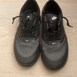 Nike Air Max Thea svart/vit sneakers - Svarta Nike Air Max Thea sneakers med vita detaljer och klassisk Swoosh på sidan. Ovandelen är i mesh och syntet för en sportig look, med snörning och platt sula. Perfekta för dig som gillar stilrena och bekväma sneakers.