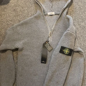 Grå stickad hoodie från Stone Island - Snygg grå stickad hoodie från Stone Island med ikonisk patch på ärmen. Tröjan har huva, lång ärm och half zip framtill. Perfekt för dig som gillar grisch och vill ha något både stilrent och bekvämt. Grovstickad känsla och riktigt skön passform. Storlek small. Jag på bilden är 171.