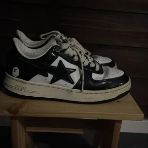 Svarta och sneakers - Säljer ett par klassisk sneakers i svart och vitt med ikonisk stjärndesign på sidan. Skorna har snörning, rund tå och platt sula. Tillverkade i glansigt syntetmaterial med Bape-logga på hälen och sidan. Perfekta för dig som gillar streetwear och vill sticka ut.