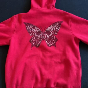 Röd hoodie med fjäril från Lecacci - Cool röd hoodie från Lecacci med glittrande stenar längs dragkedjan och en stor strassfjäril på ryggen. Tröjan har huva med snörning, ribbade muddar och är helfodrad i mjuk fleece. 