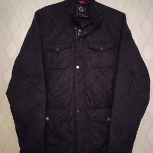 Svart quiltad jacka från Etirel - Quiltad svart jacka från Etirel Heritage Collection med snygga tryckknappar och dragkedja. Fyra fickor framtill, hög krage och diskret logga på ärmen. Perfekt för dig som gillar stilren och sportig look.