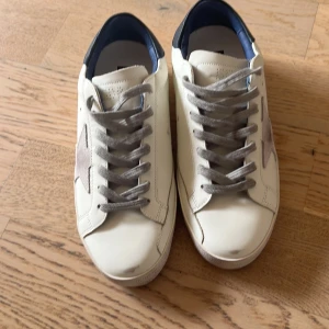 Golden Goose Superstar sneakers vit/grå - Golden Goose Superstar sneakers i vitt skinn med grå mockadetaljer och stjärna på sidan. Grå snörning, mörkblå häl och svart sula. Klassisk låg modell med rund tå och platt sula. Perfekt för dig som gillar en clean men ändå unik look.