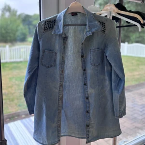 Ljusblå jeans-overshirt med nitar - Snygg ljusblå overshirt i jeansmaterial från Pink Blush. Skjortan har långa ärmar, två bröstfickor och coola svarta nitar på axlarna. Knappar hela vägen framtill och avslappnad passform. 