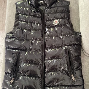 Svart väst från Moncler - Snygg svart väst från Moncler med glansig finish och klassisk logga på bröstet. Västen har två dragkedjefickor framtill och quiltad design. Perfekt att slänga över en hoodie eller tröja för en cool look.