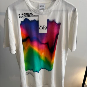 Vit t-shirt med färgstarkt tryck från Zara - Vit t-shirt från Zara med ett stort, färgstarkt grafiskt tryck på framsidan i lila, blått, rött, orange och grönt. Trycket har en cool, abstrakt vibe och textdetaljer upptill. Klassisk passform med rund hals och korta ärmar. 