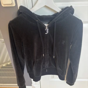 Svart velour hoodie från Juicy Couture - Svart hoodie i mjuk velour från Juicy Couture med dragkedja, huva och fickor framtill. Klassisk JC-logga broderad på bröstet och snygga detaljer i silver på snörningen. Perfekt för en chill och trendig look.