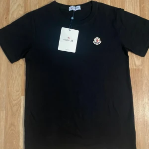 Svart Moncler t-shirt i bomull - Stilren och äkta Moncler t-shirt i svart med det ikoniska Moncler-logotypmärket på bröstet. En tidlös design som passar både till vardags och mer uppklädda tillfällen.