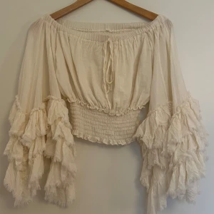 Beige volangblus med offshoulder - Supersöt beige blus med offshoulder och vida ärmar med flera lager volanger. Smockad midja och knytning framtill ger en bohemisk vibe. Perfekt för dig som gillar unika detaljer och flowy stil. Köpt i Thailand :) 