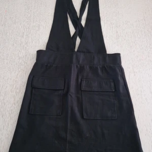 Svart hängselkjol från Name It strl 152 - Svart kort hängselkjol från Name It. Storlek 152. Stora fickor framtill och korsade axelband. Kjolen har en enkel och stilren look som passar perfekt till en cool vardagsoutfit. Mjukt och skönt material. ENDAST TESTAD!