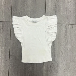 Vit ribbad topp med volangärm Zara S - Superfin vit ribbad topp från Zara i storlek S. Toppen har korta ärmar med breda volangdetaljer som ger en extra touch. Mjuk och stretchig bomullskänsla, perfekt till jeans eller kjol. Enkel men ändå unik med sina volanger på axlarna.