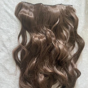 Brun hårförlängning med clips - Säljer en vågig hårförlängning i mellanbrun nyans med clips. Håret är syntetiskt och har en naturlig glans. Fästet är gjort av svart spets med flera clips för enkel applicering. Perfekt för att snabbt få längre och fylligare hår. Helt ny 