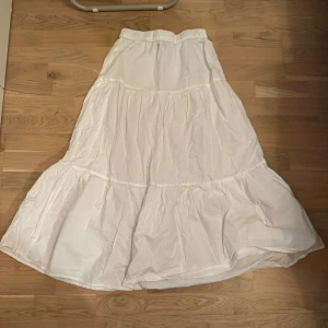 Vit maxikjol från Gina Tricot - Superfin vit långkjol från Gina Tricot med volangiga lager och resår i midjan. Kjolen är luftig och har en flowy känsla, perfekt för varma dagar. 