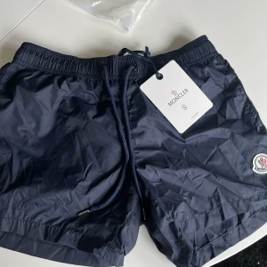 Mörkblå Moncler badshorts med snörning - Snygga mörkblå badshorts från Moncler med klassisk logga på benet. Shortsen har elastisk midja med snörning, meshfoder och en bakficka med dragkedja och röd/vit detalj. Perfekta för stranden eller poolen, riktigt stilrena och sportiga.