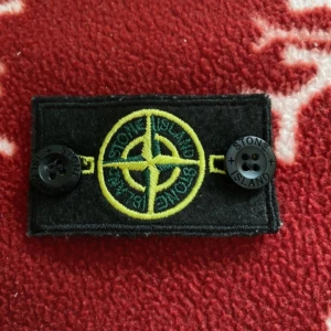 Stone Island svart patch med logga - Svart patch från Stone Island med den ikoniska kompassloggan broderad i gult och grönt. Två svarta knappar med Stone Island-tryck på sidorna. Perfekt att fästa på jacka eller tröja för en streetwear-look.