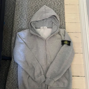 Grå hoodie från Stone Island - Snygg grå hoodie från Stone Island med dragkedja och klassisk logga på ärmen. Tröjan har huva, ribbade muddar och fickor framtill. Perfekt för en chill och stilren look.