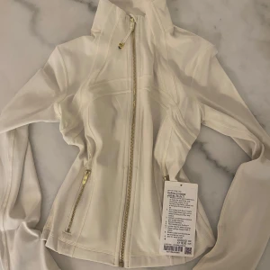 Vit Define Cropped jacka från Lululemon - Säljer en vit Define Cropped jacka från Lululemon i mjukt Nulu-material med guldfärgade dragkedjor. Jackan har slim fit, långa ärmar med tumhål och praktiska fickor med dragkedja. Perfekt för dig som gillar sportig och clean stil.