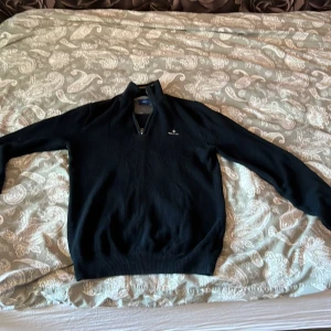 Svart stickad half zip-tröja GANT - Svart stickad tröja från GANT med half zip och hög krage. Klassisk och stilren modell med diskret logga på bröstet. Långärmad och ribbade muddar vid ärmslut och nederkant. Perfekt för lager-på-lager under höst och vinter.