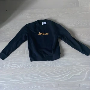 Svart sweatshirt från Axel Arigato - Svart sweatshirt från Axel Arigato med gul broderad text 'Arigato' på bröstet. Klassisk rund halsringning, ribbade muddar och lång ärm. Perfekt för en clean och stilren look. Materialet känns mjukt och skönt mot huden.