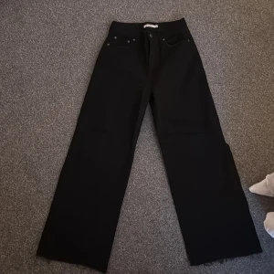 Svarta wide jeans från NA-KD, stl 38 - Säljer ett par svarta jeans från NA-KD i storlek 38. Byxorna har hög midja, raka vida ben och klassisk femficksdesign. Snygga nitar vid fickorna och stilren look som passar till allt. Materialet är kraftigt och håller formen bra.